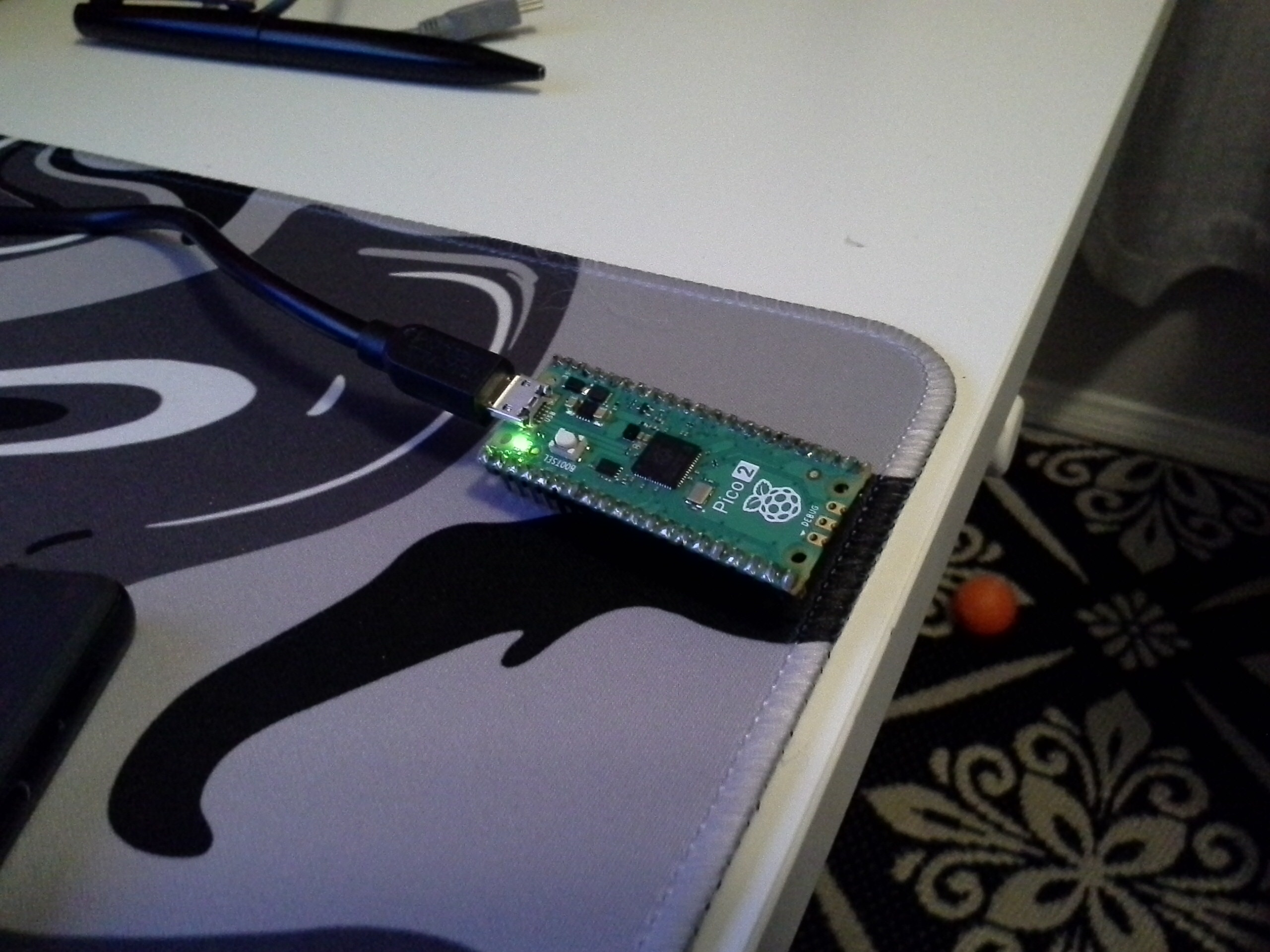 rasberry pi pico on a desk mat conected to a computer with its light on.